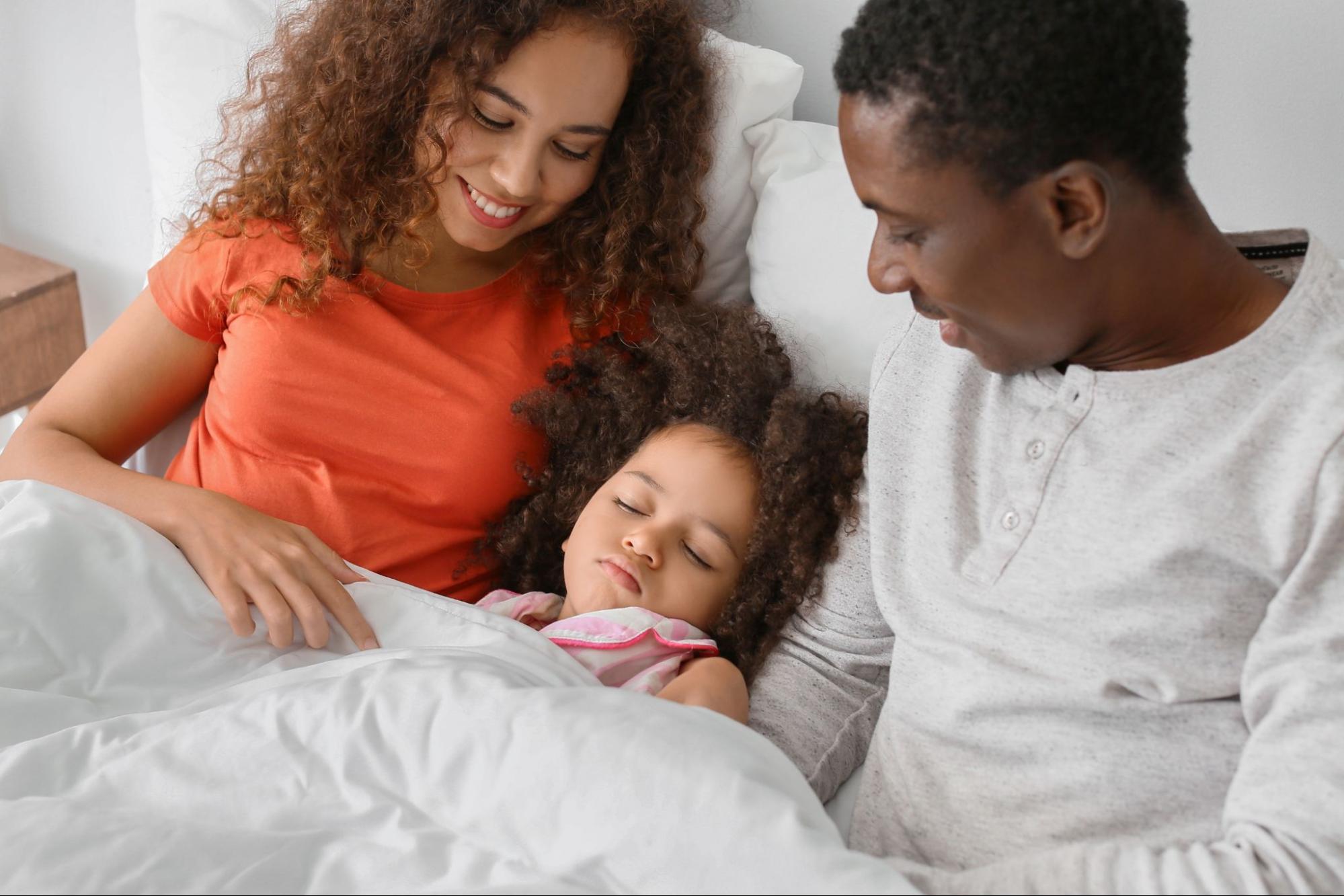 A woman and man sit on a bed, smiling at a young child who is asleep under a white blanket